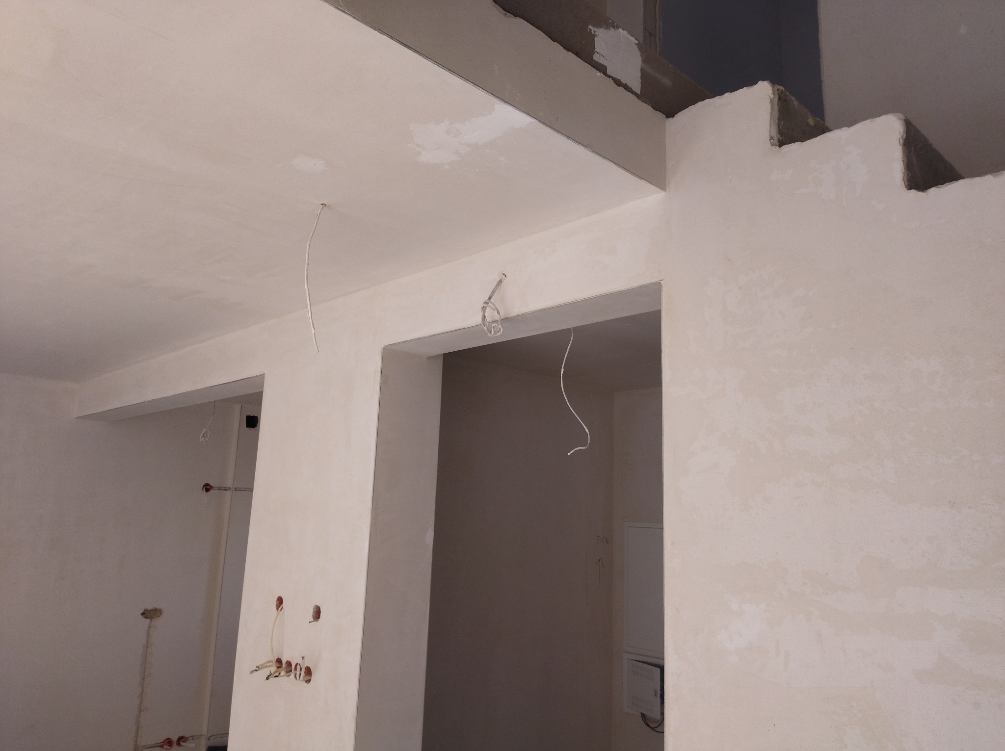 Wnętrze w trakcie prac wykończeniowych: widoczne przewody elektryczne wystające z sufitu i ścian, przygotowane pod montaż osprzętu. Ściany otynkowane, sufit pomalowany.