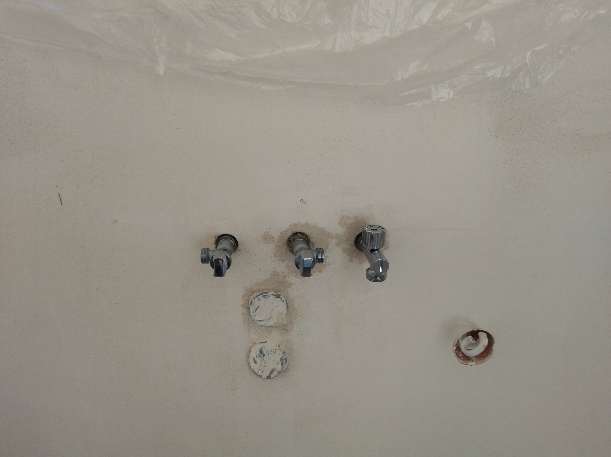Ściana z trzema zaworami wodnymi i otworami, widoczne ślady po instalacji. U góry folia ochronna, co sugeruje prace remontowe.