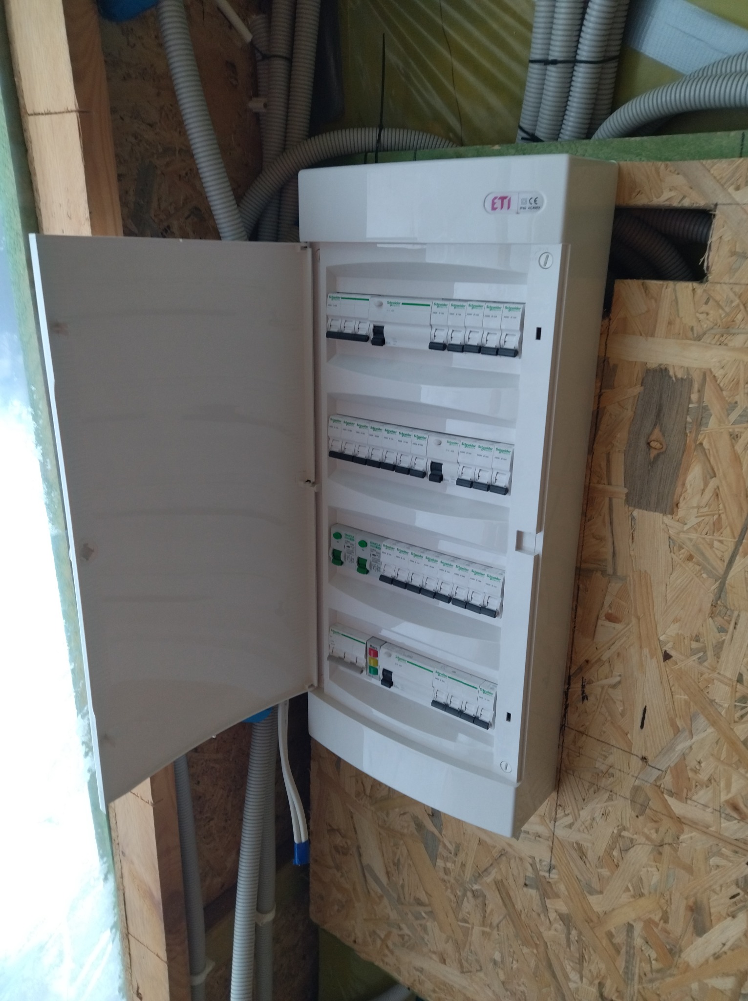 Otwarta skrzynka elektryczna ETI z bezpiecznikami na ścianie z płyty OSB, widoczne kable w peszlach. Instalacja w trakcie realizacji.