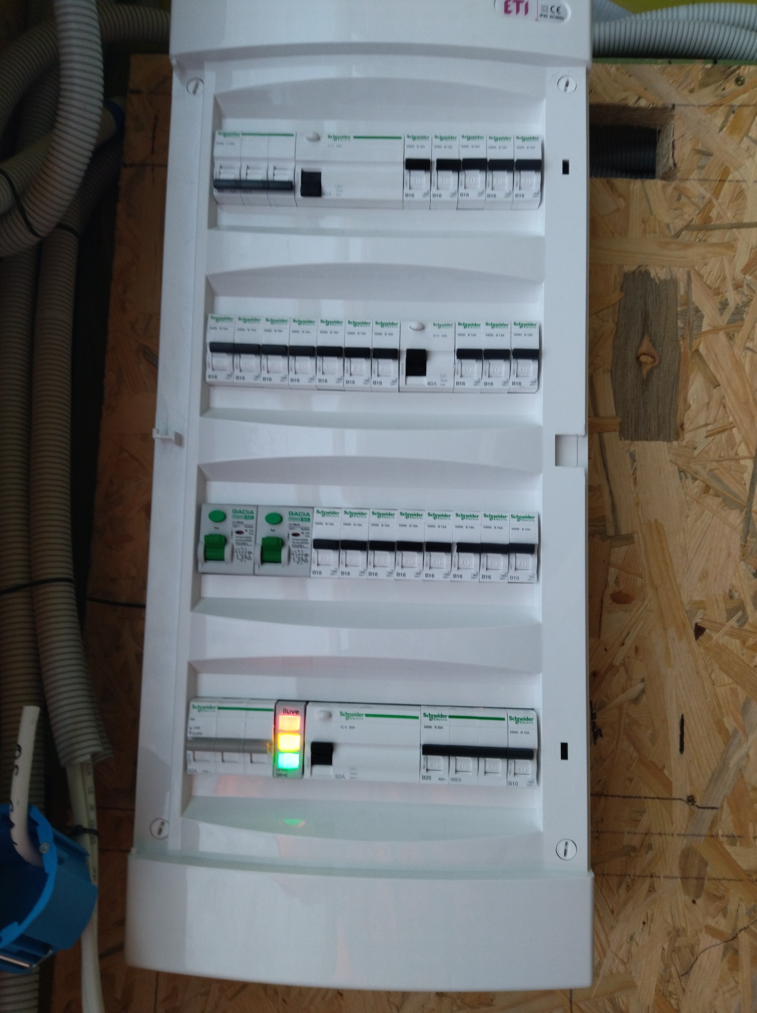 Rozdzielnica elektryczna Schneider z bezpiecznikami i wyłącznikami, zamontowana na ścianie z płyty OSB. Widoczne przewody w peszlu i elementy instalacji.