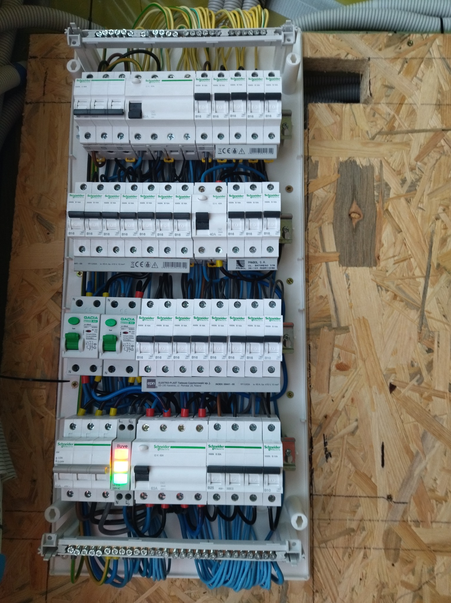 Rozdzielnica elektryczna z bezpiecznikami i okablowaniem, zamontowana na płycie OSB. Widoczne elementy marki Schneider Electric i Gacia.