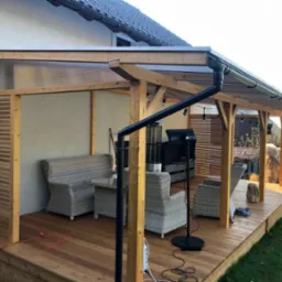 Zadaszony taras drewniany z meblami ogrodowymi z rattanu, oświetleniem i grillem, widok na ogród.