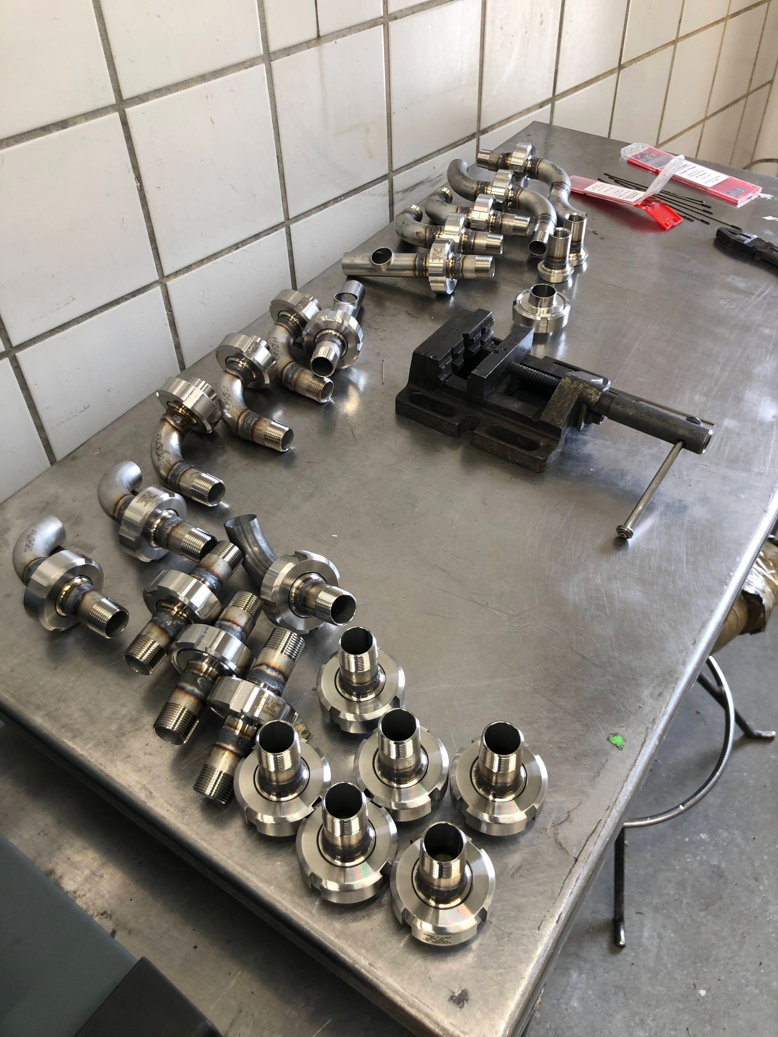 Metalowe złączki rurowe, niektóre z przyspawanymi kolankami, leżące na metalowym stole warsztatowym obok imadła i elektrod spawalniczych na tle ściany z białych płytek.