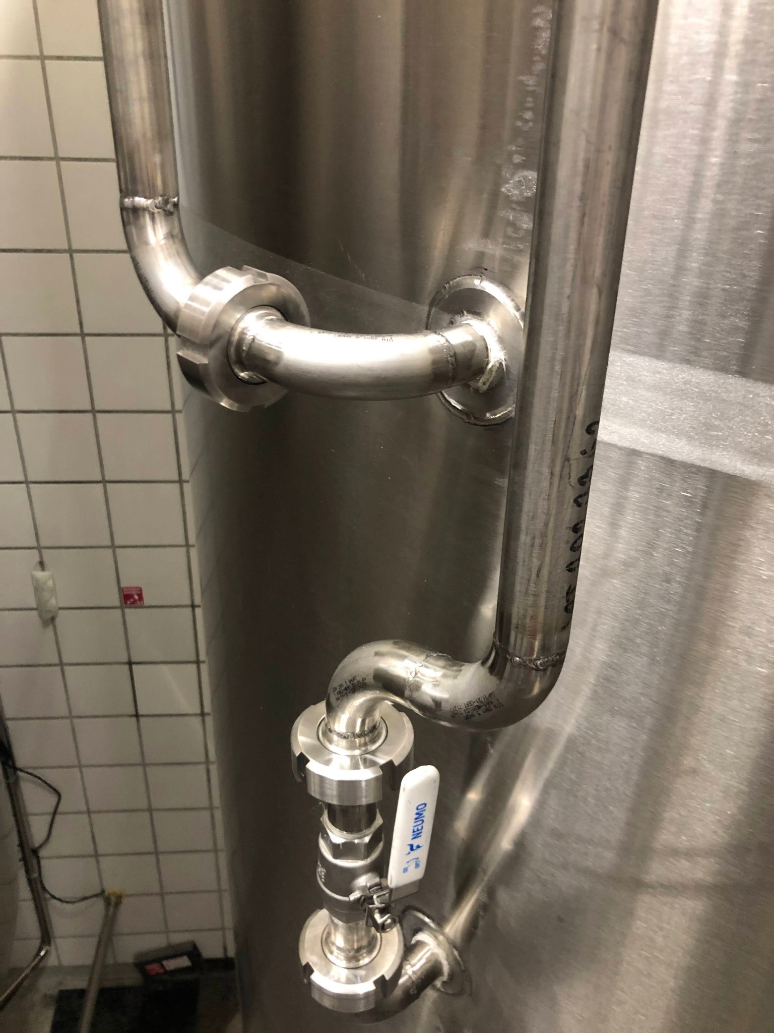 Metalowy zbiornik z rurami i zaworem marki NEUMO, widoczne spawy i oznaczenia na rurach, tło z płytek ceramicznych.