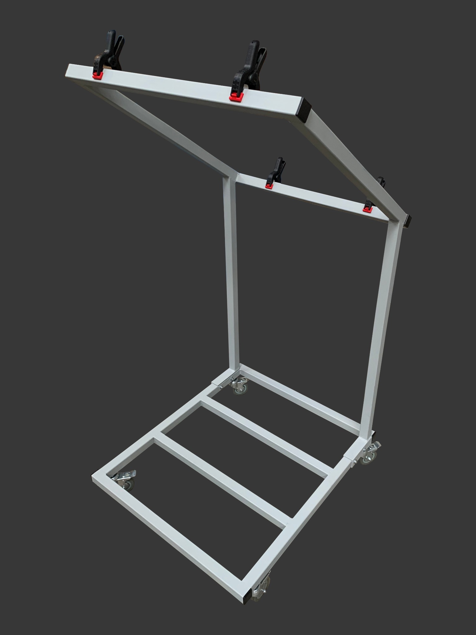 Metalowa konstrukcja z kółkami, prawdopodobnie element warsztatowy, z uchwytami zaciskowymi, pomalowana na biało, na szarym tle.