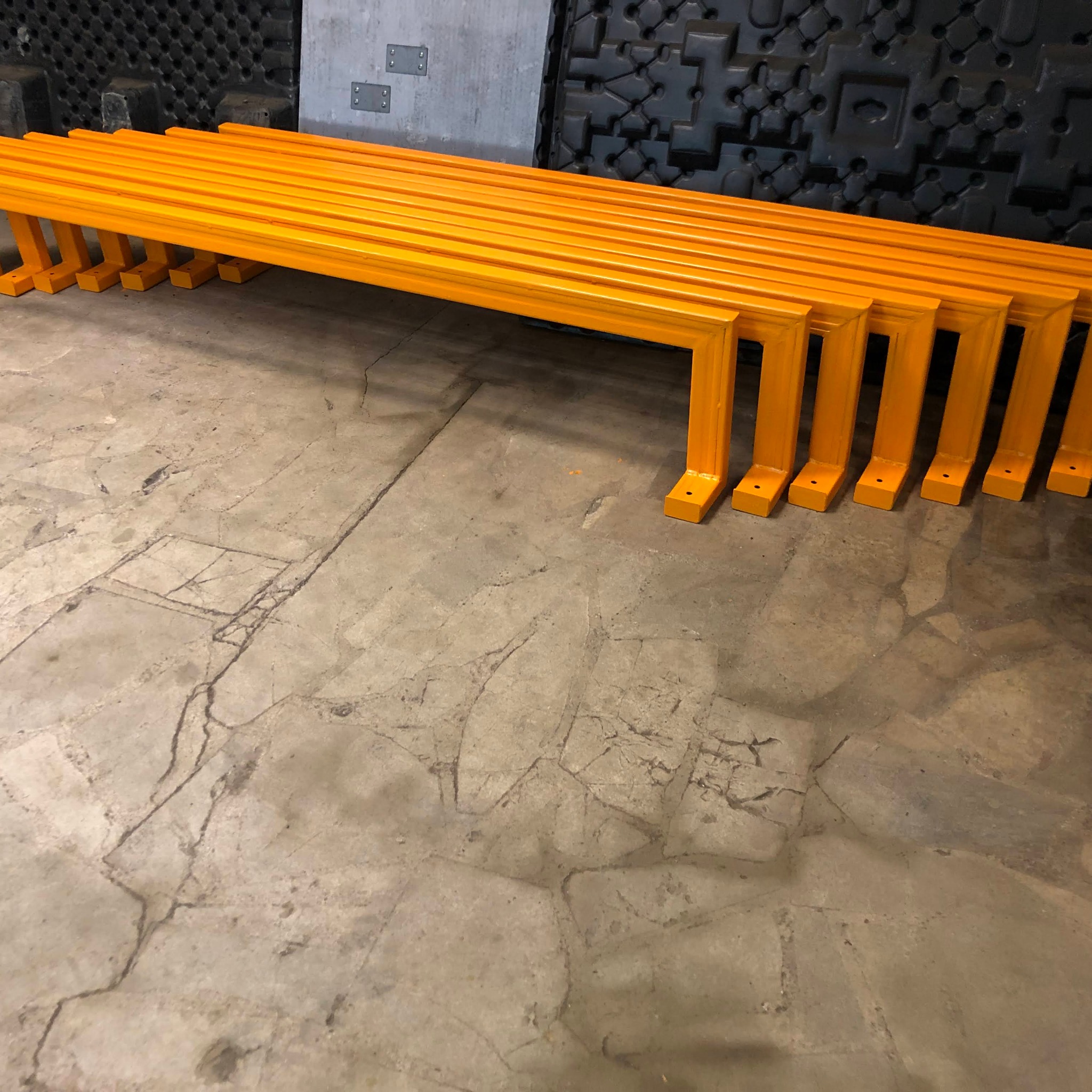 Rząd pomarańczowych, metalowych konstrukcji ramowych z otworami na śruby, ułożonych na betonowej podłodze w Bielsku-Białej, prawdopodobnie w warsztacie.