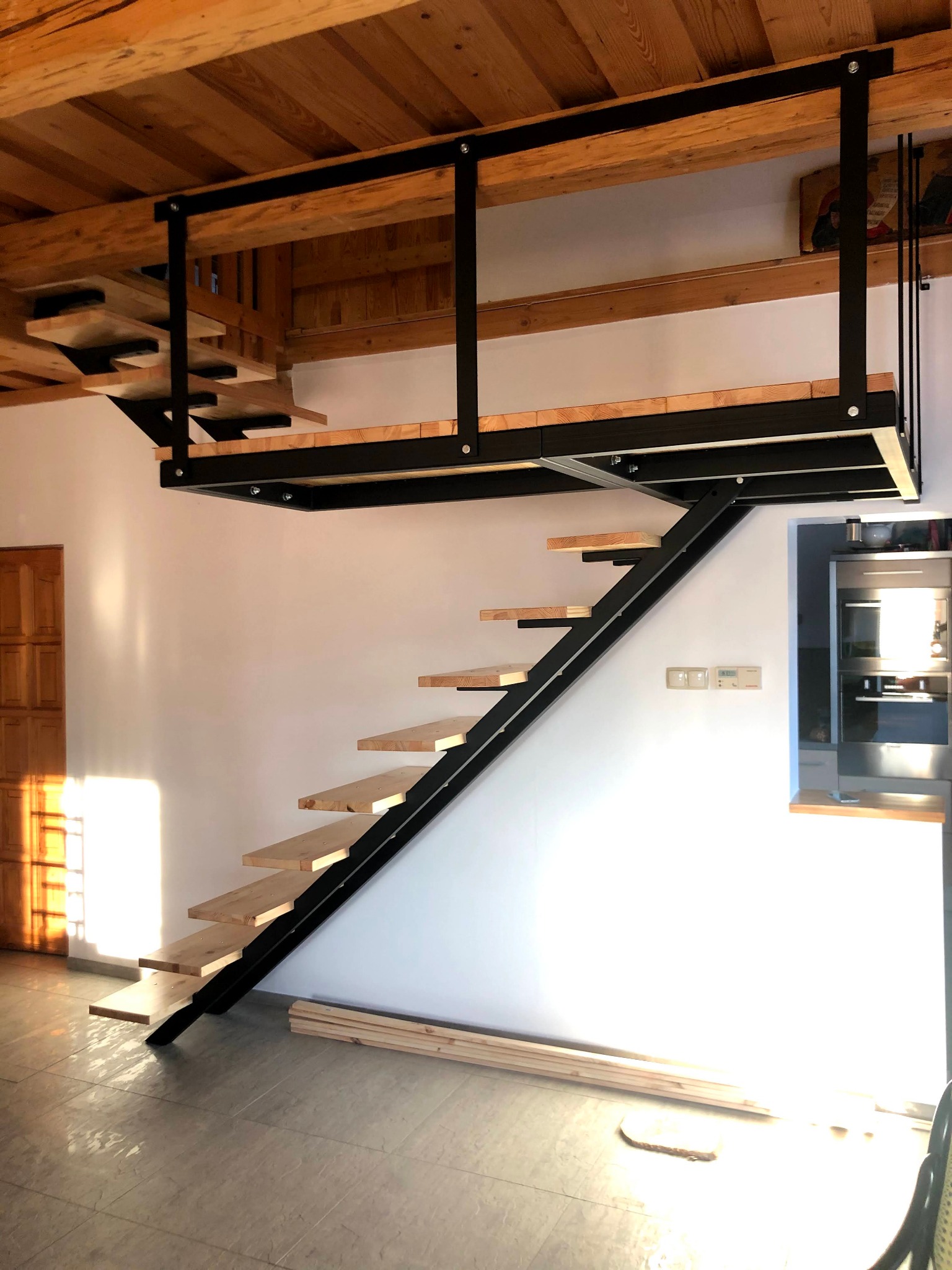 Nowoczesne schody z czarną metalową konstrukcją i drewnianymi stopniami prowadzące na antresolę, widoczne surowe drewno na podłodze i drewniany sufit.