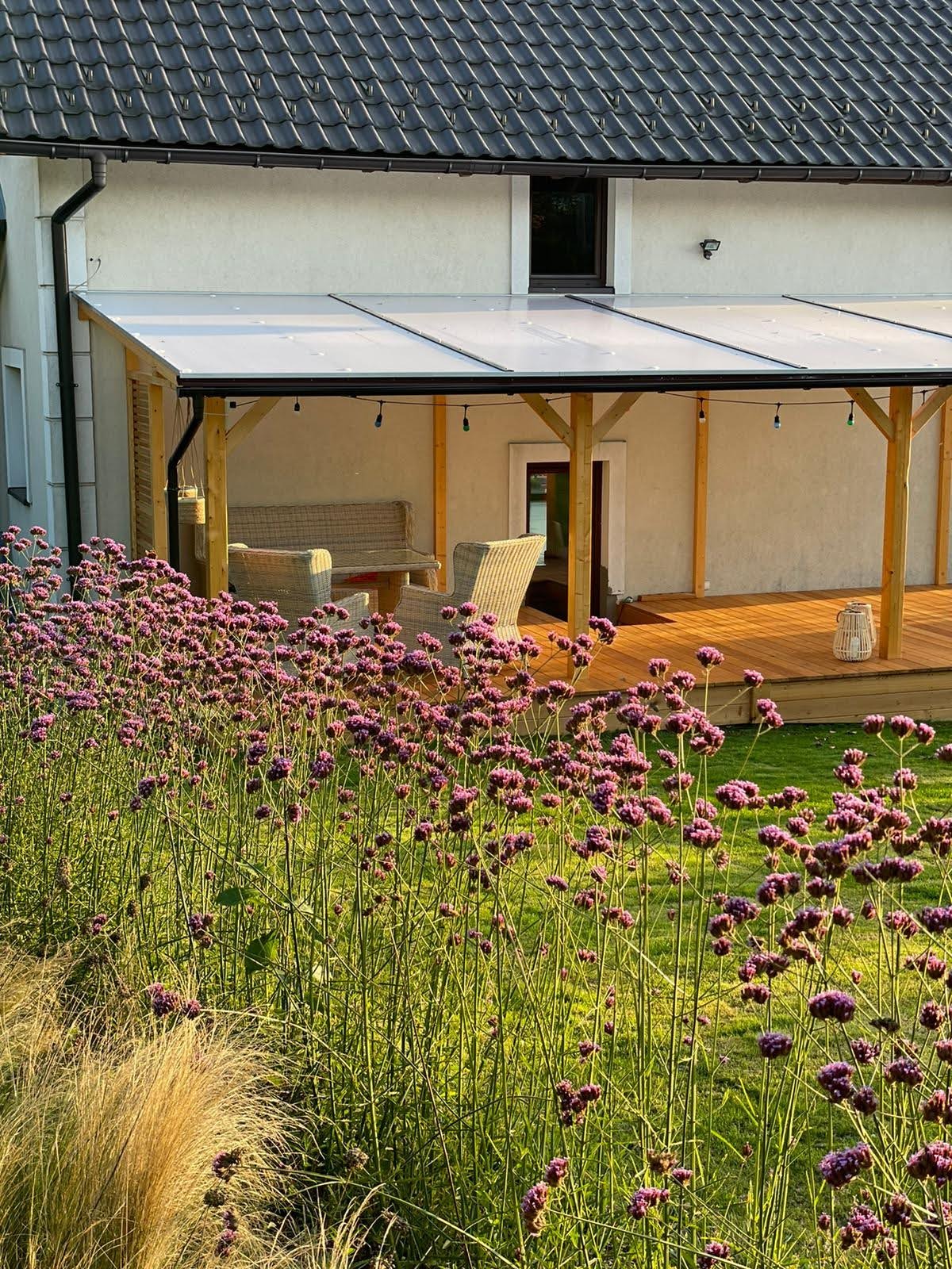 Zadaszony taras z drewnianą podłogą, wiklinowymi meblami i dekoracyjnym oświetleniem, widziany zza kwitnących na fioletowo kwiatów w ogrodzie.