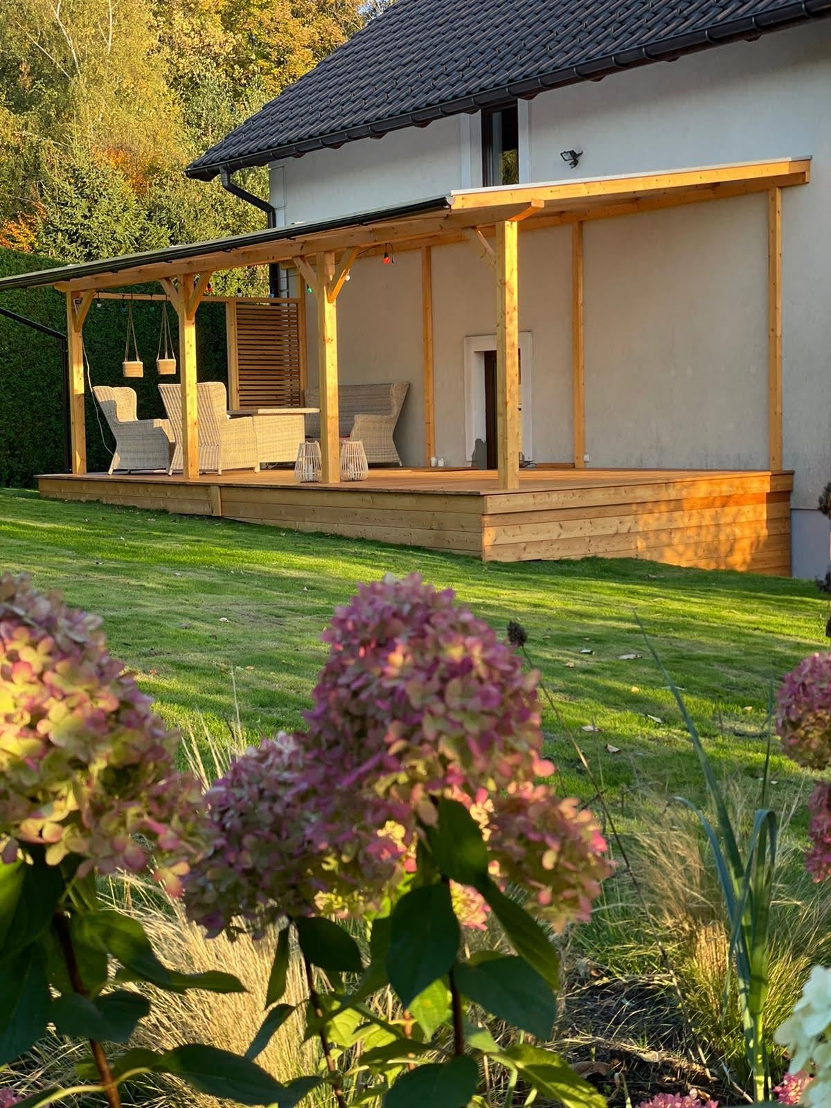 Zadaszony drewniany taras przy domu z meblami ogrodowymi z rattanu i wiszącymi lampionami, z perspektywy ogrodu z kwitnącymi hortensjami w słoneczny dzień.