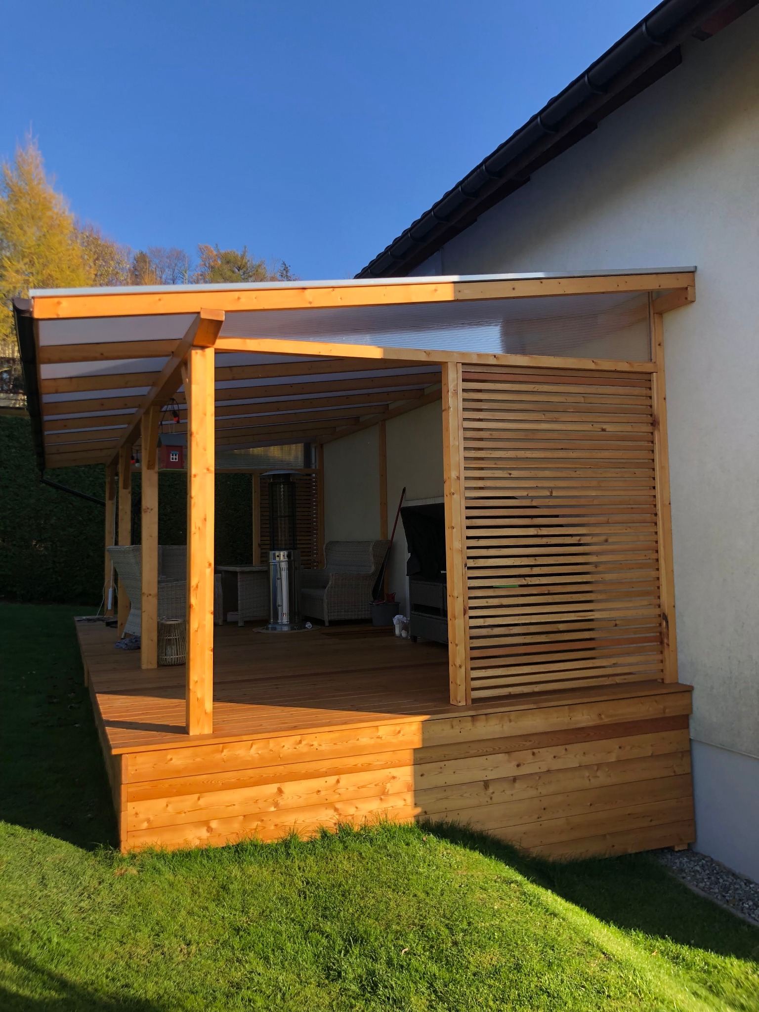 Zadaszony taras z drewna z ażurową ścianką, oświetlony popołudniowym słońcem, z widocznym wyposażeniem wypoczynkowym i grzejnikiem.