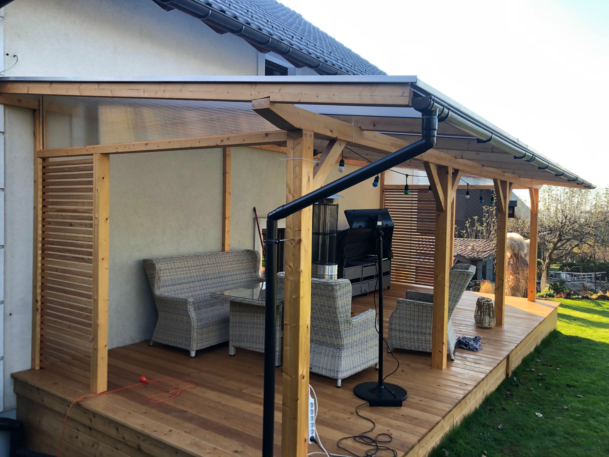 Zadaszony taras drewniany z meblami ogrodowymi z rattanu, oświetleniem i grillem, widok na ogród.