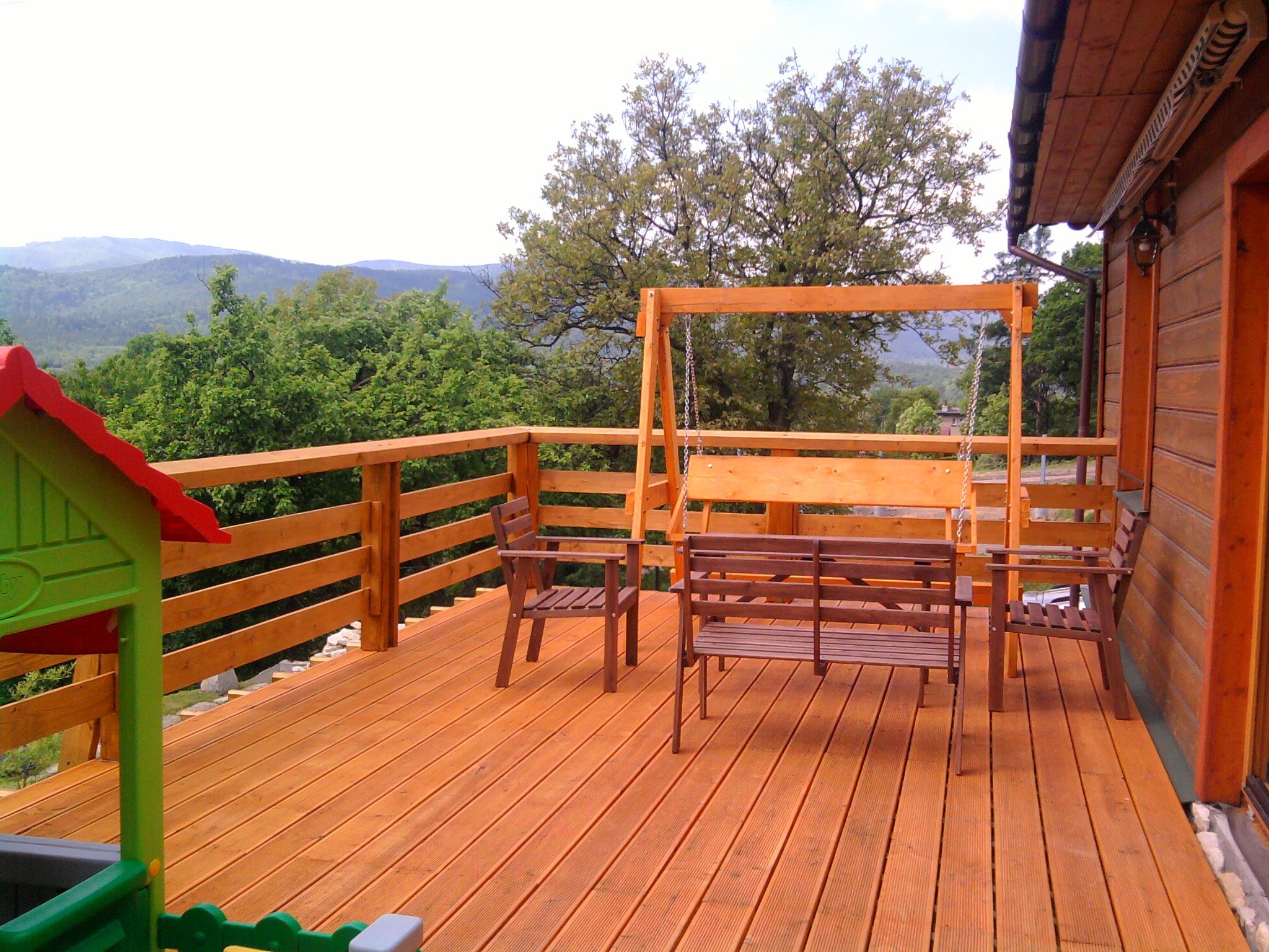 Drewniany taras z widokiem na góry, wyposażony w huśtawkę, ławkę i krzesła, z elementami placu zabaw dla dzieci w rogu.