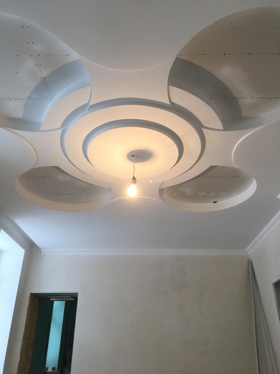 Dekoracyjny sufit z płyty karton gips z okrągłymi elementami i centralnym oświetleniem w surowym wnętrzu. Widoczne ślady po szpachlowaniu.