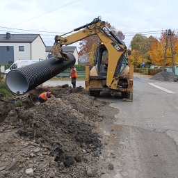 Usługi koparko-ładowarką Szymala Grzegorz - Żółta koparko-ładowarka CAT umieszcza dużą, czarną rurę w wykopie na poboczu drogi, pracownicy w pomarańczowych kamizelkach kontrolują proces.