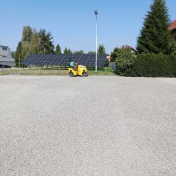 Usługi koparko-ładowarką Szymala Grzegorz - Operator na żółtym walcu drogowym zagęszcza szarą nawierzchnię z kruszywa na tle paneli słonecznych i zieleni.