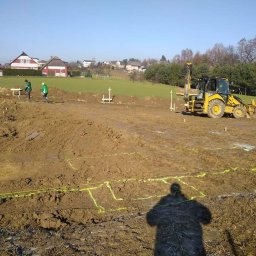 Usługi koparko-ładowarką Szymala Grzegorz - Żółta koparka z wiertnicą na placu budowy, wytyczone fundamenty budynku, dwóch pracowników przy pomiarach, cień fotografa w dolnej części kadru.