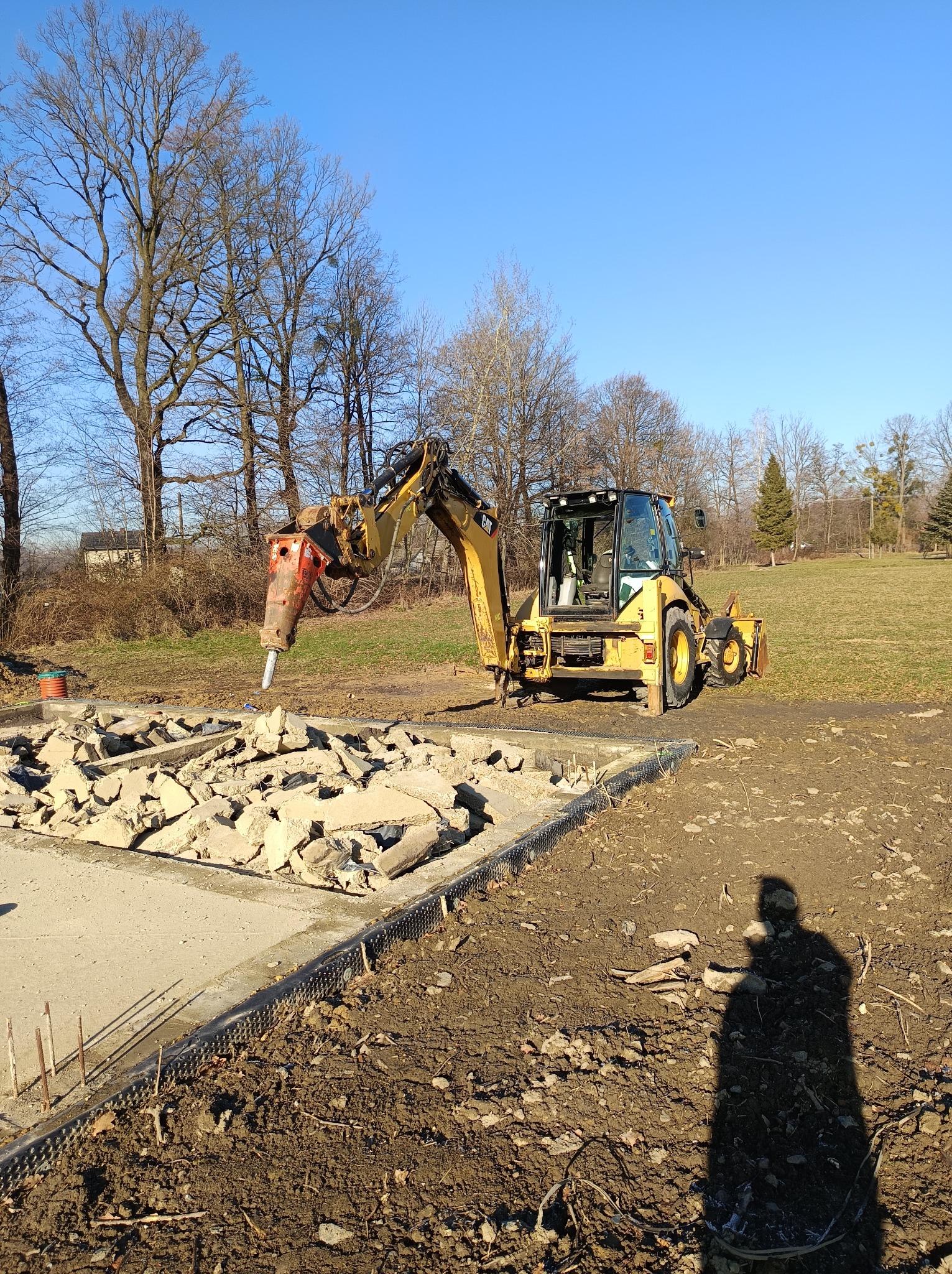 Żółta koparko-ładowarka z młotem hydraulicznym kruszy betonową płytę fundamentową na placu budowy w słoneczny dzień.