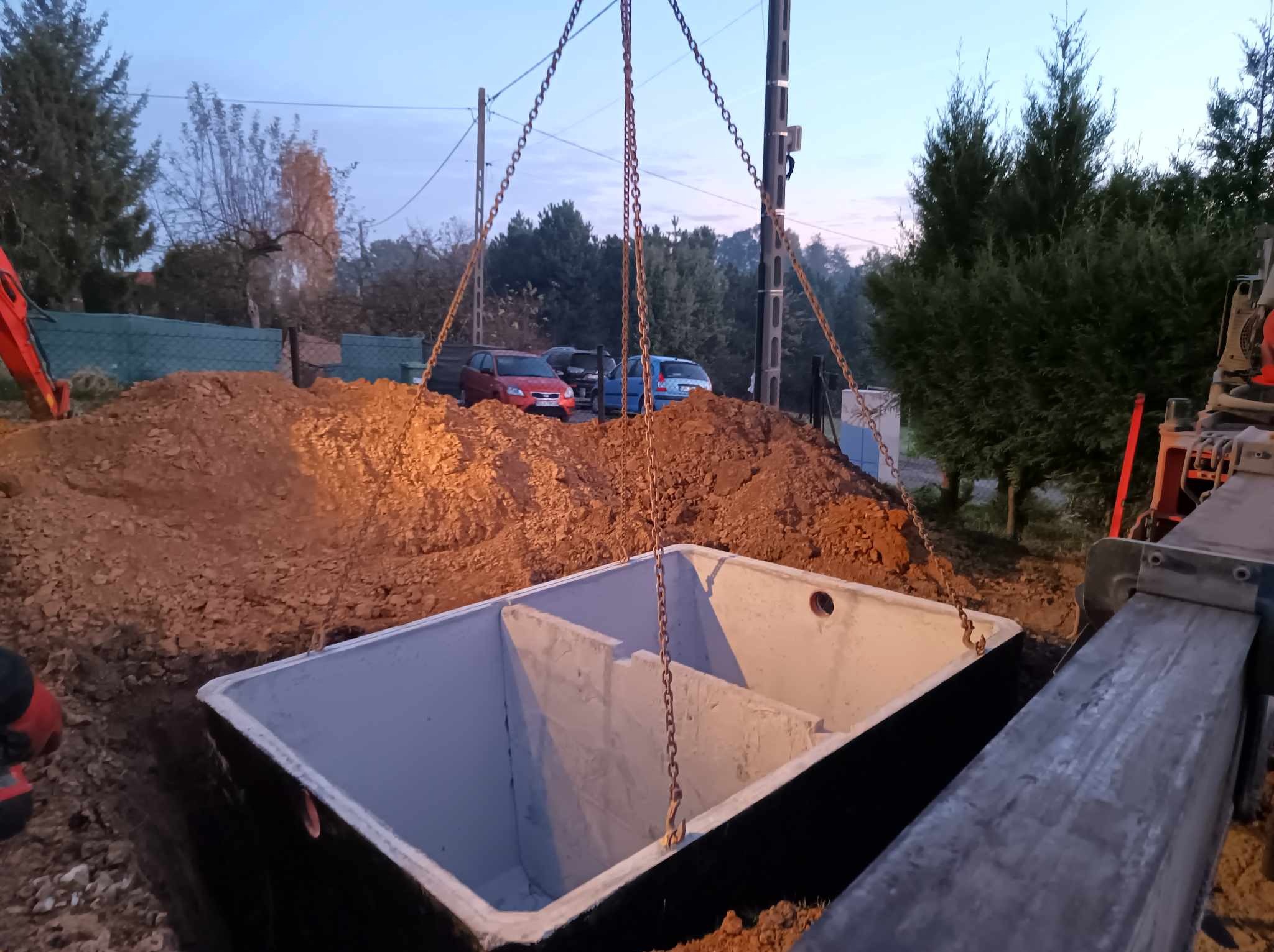Betonowy zbiornik wielokomorowy, zawieszony na łańcuchach nad wykopem, w tle widoczna sterta ziemi, częściowo widoczna koparka i zabudowa mieszkalna