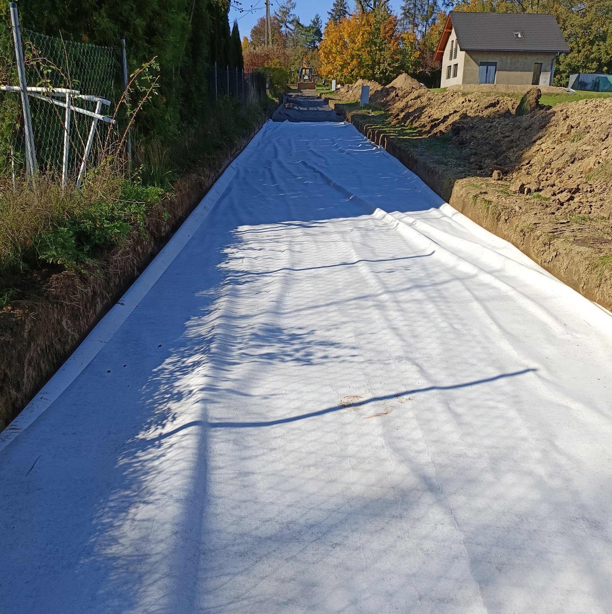 Szeroki wykop w ziemi wyłożony białą geowłókniną, w tle widoczna koparka i budynek mieszkalny.