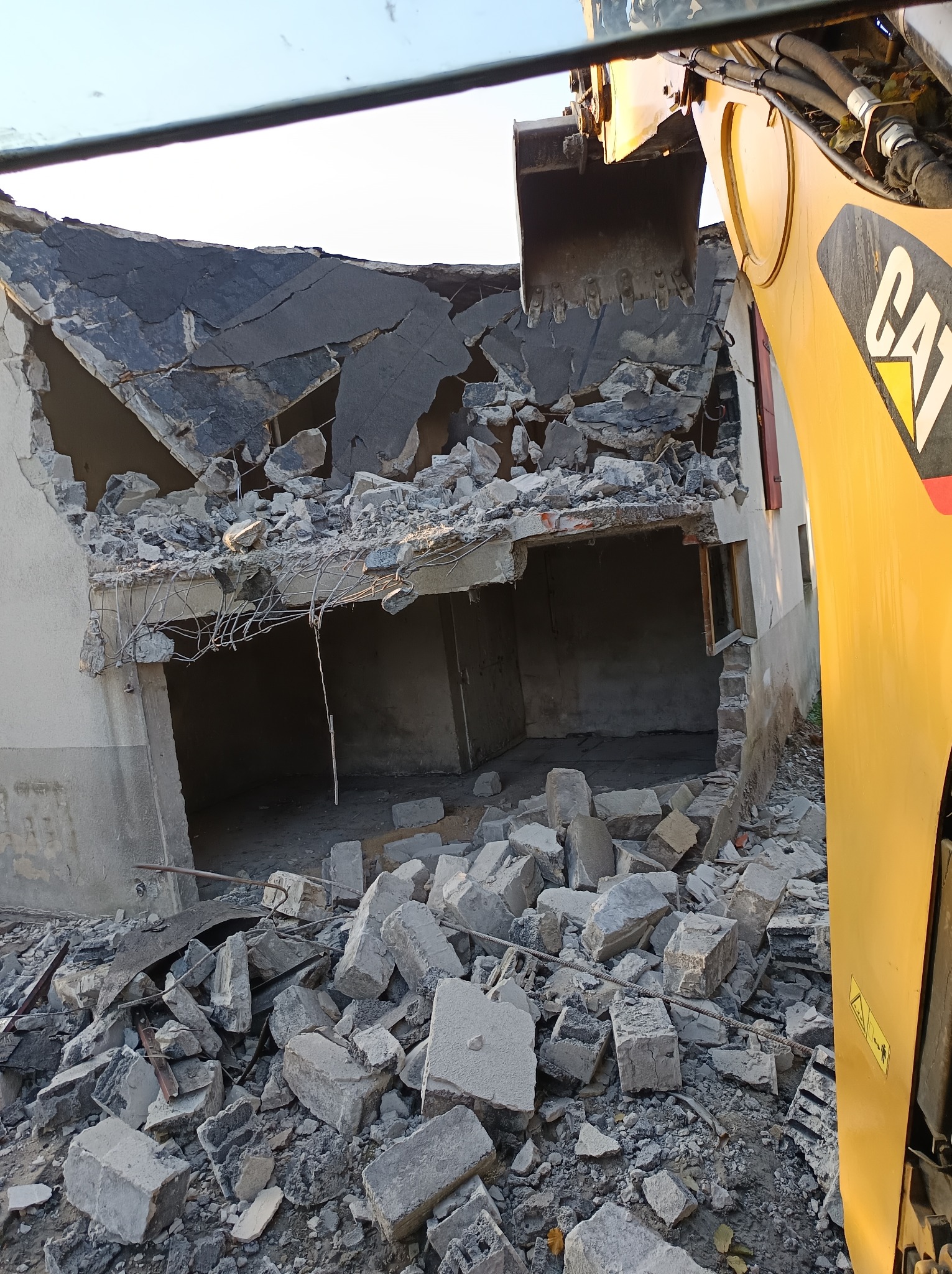 Żółta koparka CAT z łyżką na tle zrujnowanego budynku z widocznymi elementami konstrukcyjnymi i stertą gruzu.