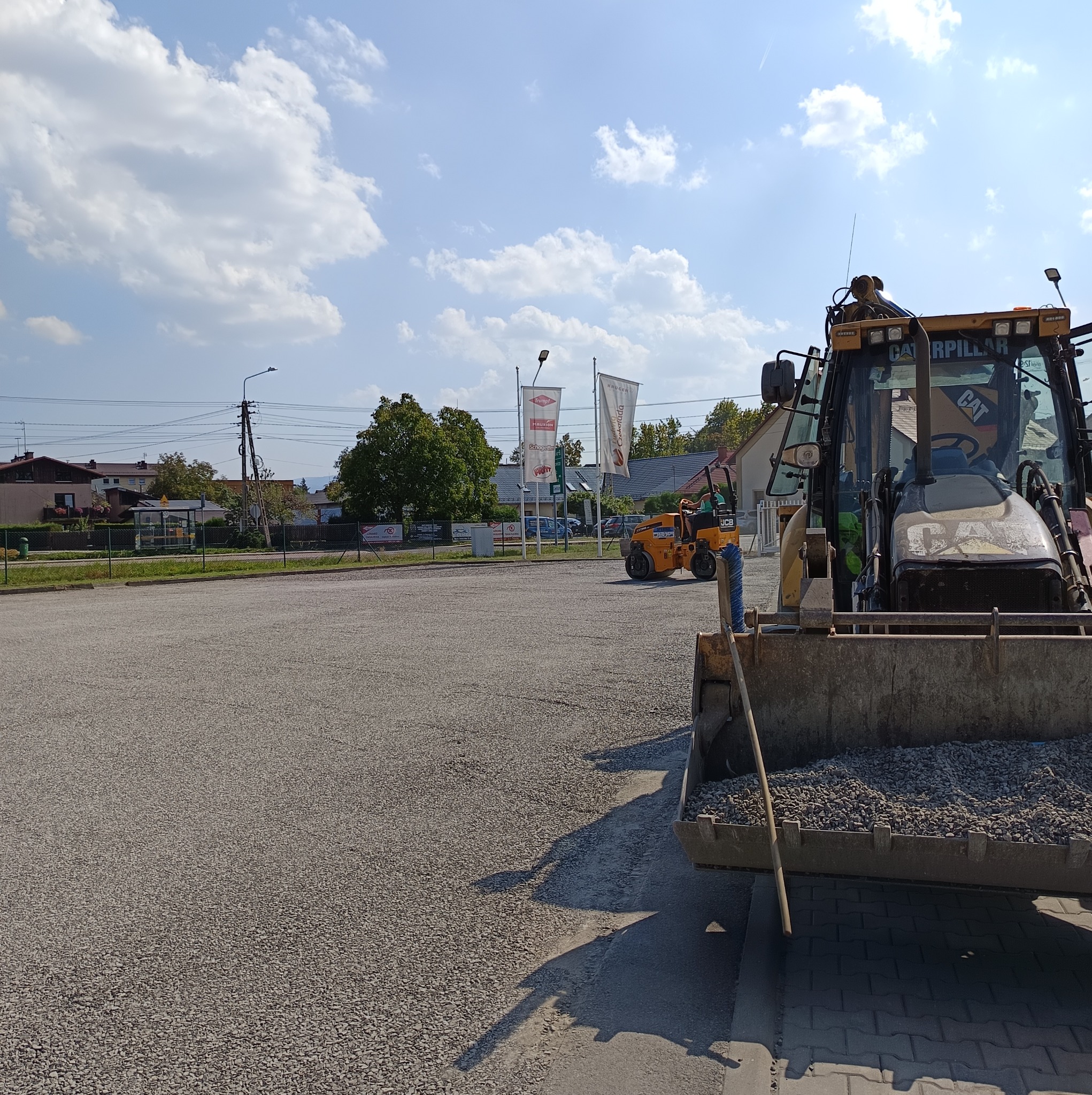 Żwirowa nawierzchnia placu budowy z ładowarką CAT i walcem drogowym JCB w tle, widok w słoneczny dzień.