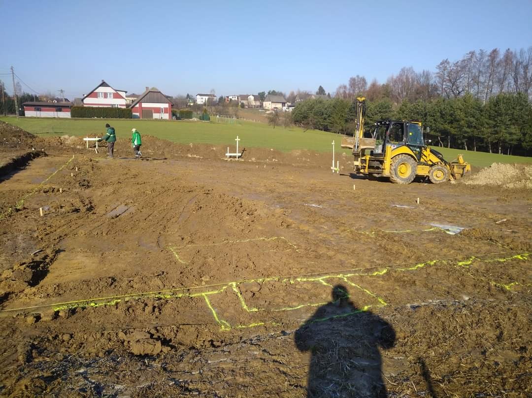 Żółta koparka z wiertnicą na placu budowy, wytyczone fundamenty budynku, dwóch pracowników przy pomiarach, cień fotografa w dolnej części kadru.