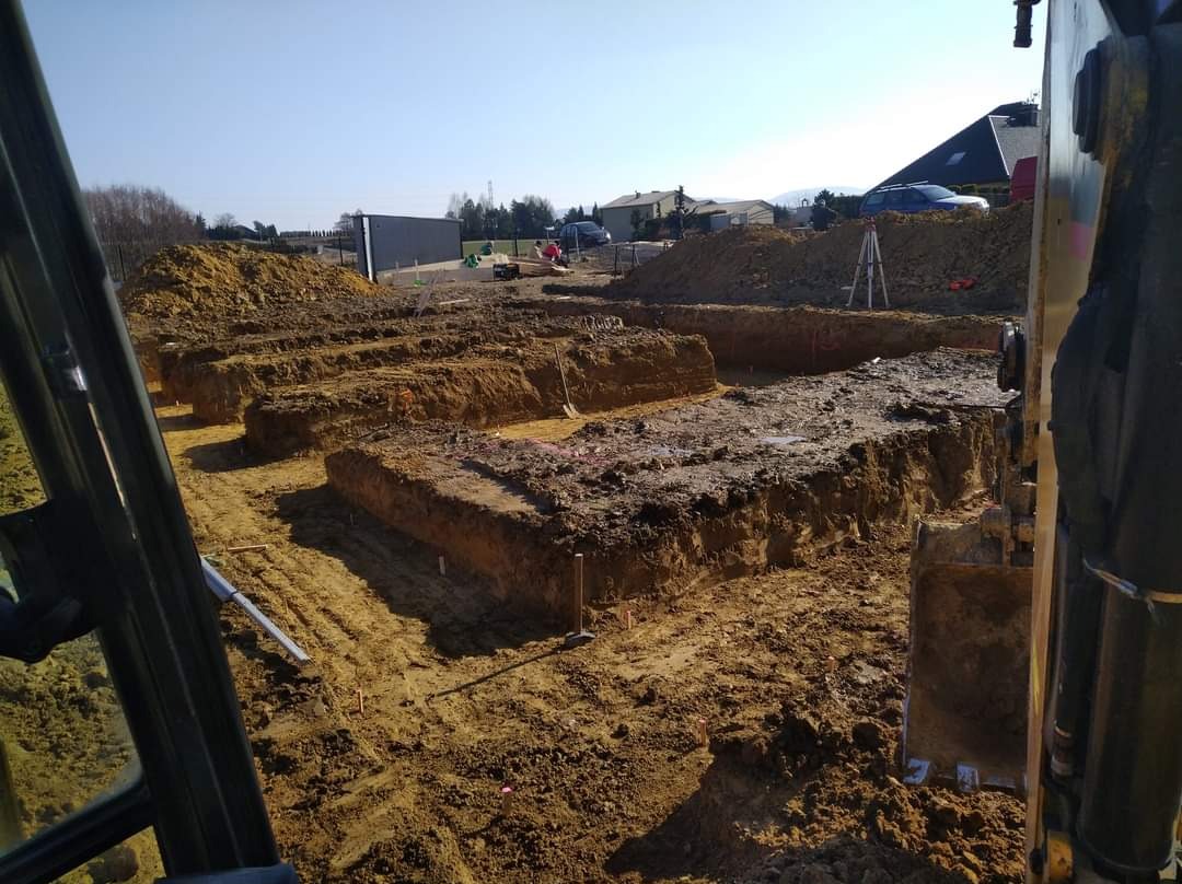 Wykopy pod fundamenty z widoczną ziemią, palikami wyznaczającymi granice oraz fragmentem kabiny koparki po prawej stronie.