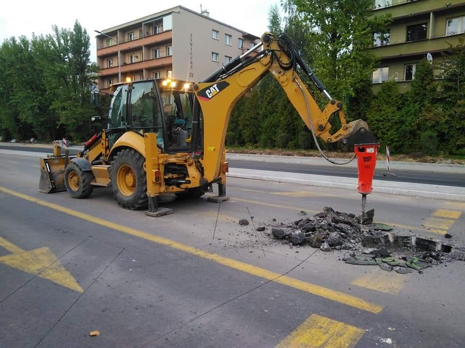 Żółta koparko-ładowarka CAT z włączonymi światłami roboczymi, rozbijająca asfalt na jezdni za pomocą czerwonego młota pneumatycznego, z widocznymi odłamkami i strzałką kierunkową na asfalcie.