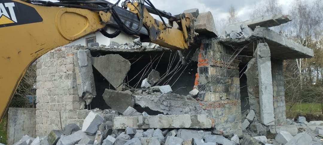 Żółta koparka CAT kruszy resztki murowanego budynku z cegieł i betonu, widoczne wystające druty zbrojeniowe i gruzowisko u podstawy.