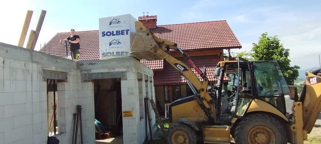 Żółta koparko-ładowarka CAT 432E podnosi paletę bloczków Solbet na budowę domu jednorodzinnego, na górze konstrukcji stoi pracownik w czarnej koszulce.