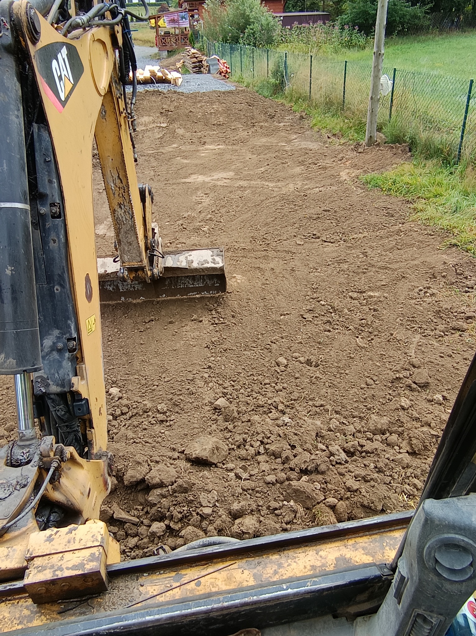 Żółta koparka CAT wyrównuje teren pod budowę drogi na działce z zielonym ogrodzeniem z siatki.