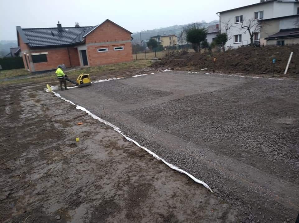 Pracownik w żółtej kurtce zagęszcza gruz wibratorem na budowie domu jednorodzinnego, widoczne białe taśmy wyznaczające obszar prac.