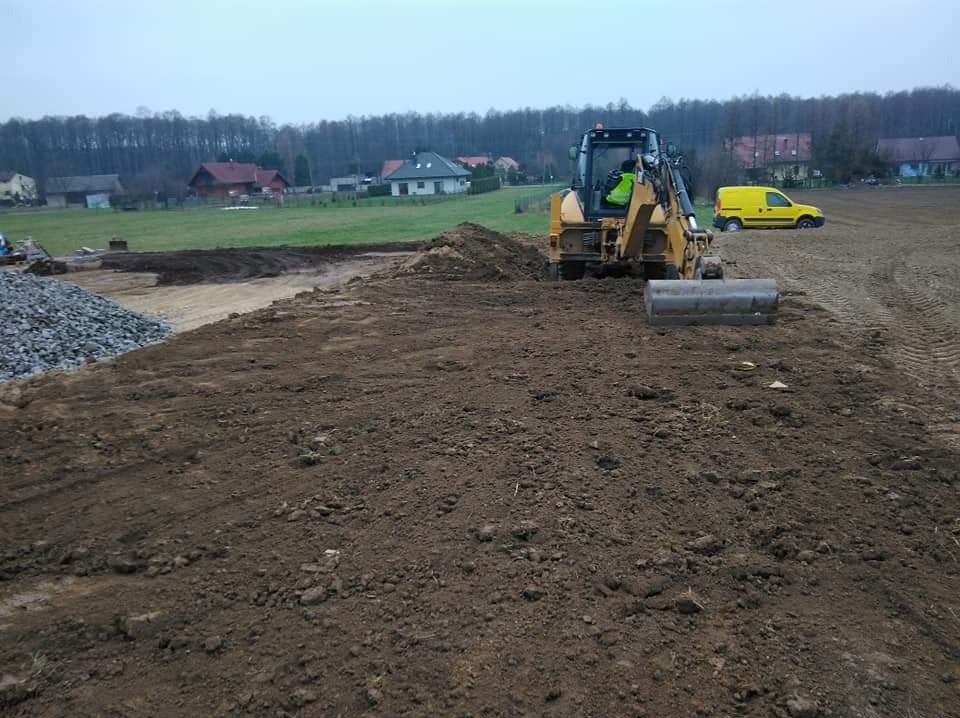 Żółta koparko-ładowarka wyrównuje ziemię na placu budowy, widoczny stos kamieni i żółty samochód dostawczy w tle.