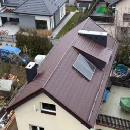 Widok z góry na dom z brązowym dachem z dachówki, z zamontowanymi panelami słonecznymi i nowym kominem. Na podwórku zaparkowany niebieski bus firmy dekarskiej.