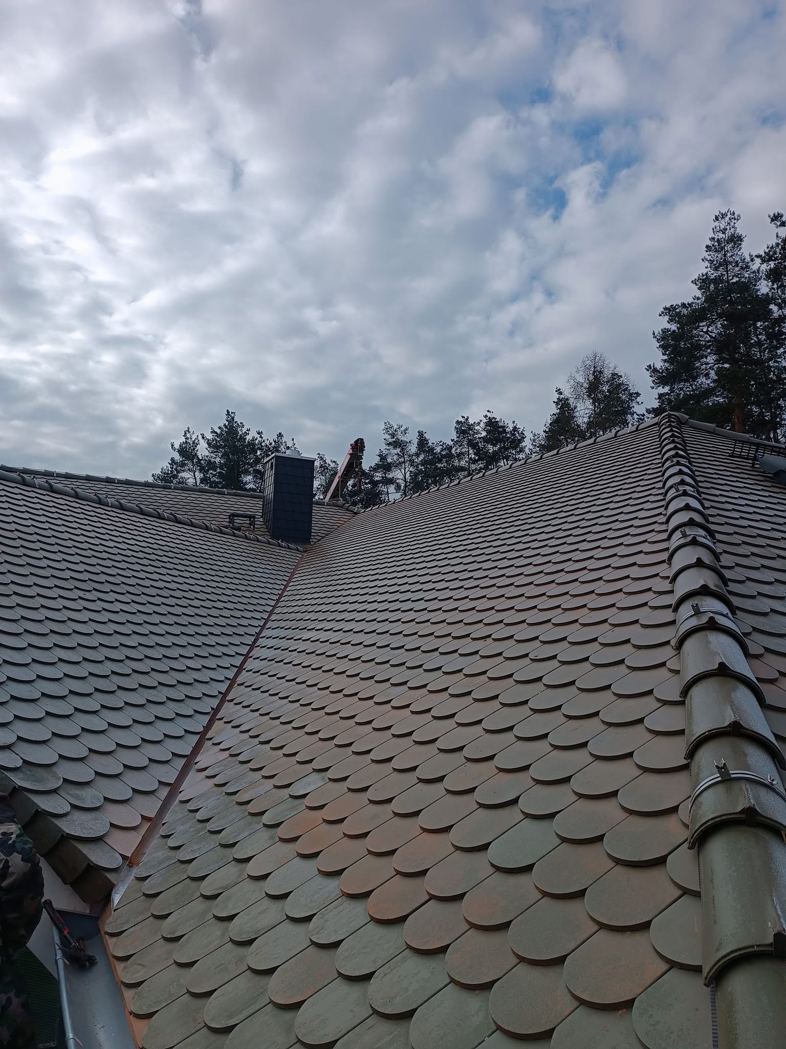 Dach pokryty łuską ceramiczną w odcieniach szarości, widoczny komin i fragment montowanej rynny, na tle pochmurnego nieba i drzew.