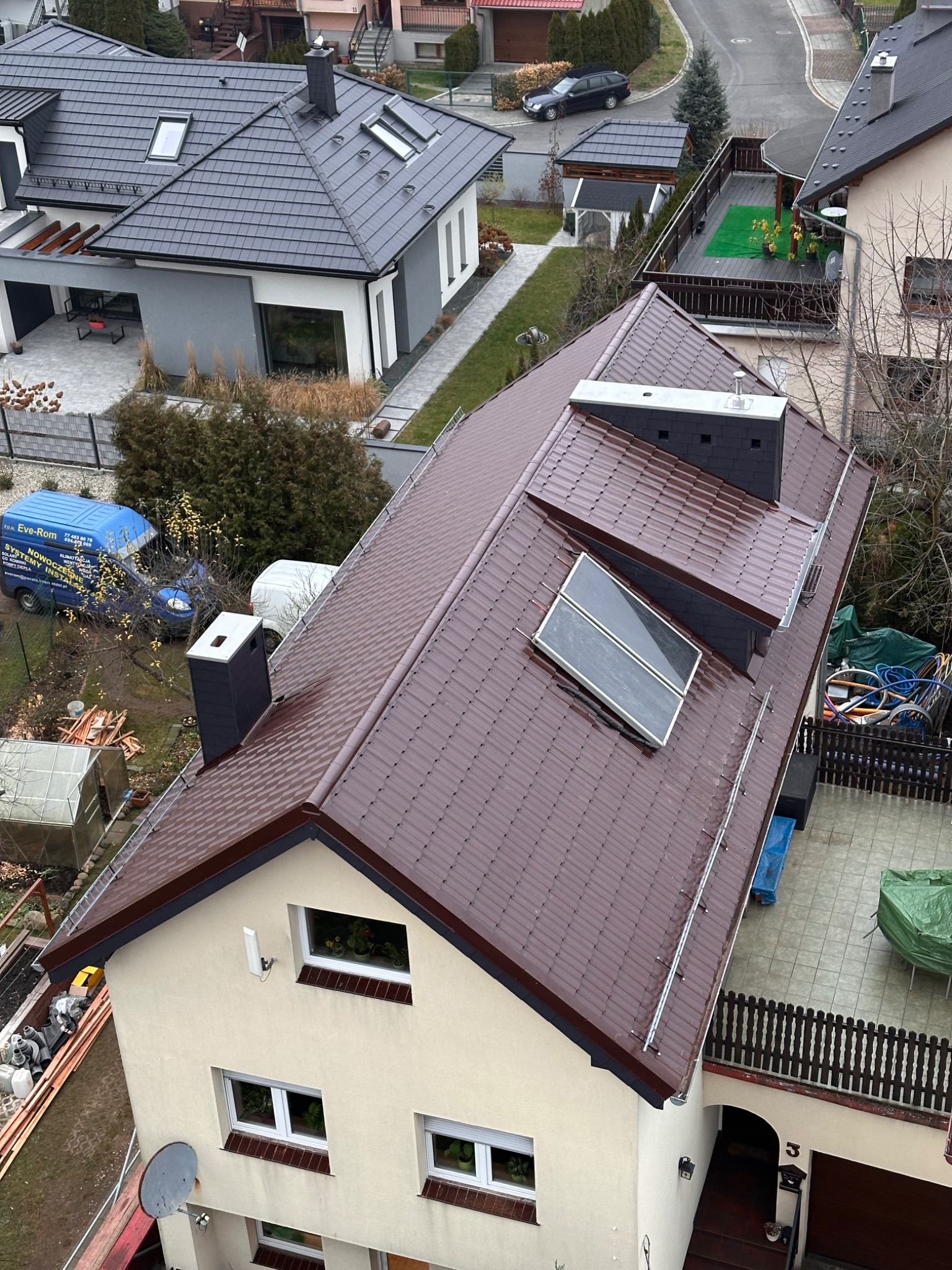 Widok z góry na dom z brązowym dachem z dachówki, z zamontowanymi panelami słonecznymi i nowym kominem. Na podwórku zaparkowany niebieski bus firmy dekarskiej.