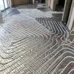 USŁUGI INSTALACYJNE KRYSTIAN SŁOWIOK - Rozłożona instalacja ogrzewania podłogowego na folii aluminiowej z siatką, widoczne białe rurki w regularnych odstępach, surowy stan wykończenia wnętrza.