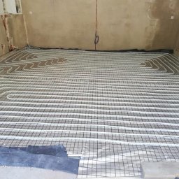 USŁUGI INSTALACYJNE KRYSTIAN SŁOWIOK - Instalacja ogrzewania podłogowego w surowym wnętrzu, rury ułożone na siatce z folią aluminiową, widoczne fragmenty betonu i instalacji elektrycznej.