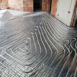 USŁUGI INSTALACYJNE KRYSTIAN SŁOWIOK - Rozłożone rury ogrzewania podłogowego na srebrnej folii izolacyjnej z siatką, surowy stan wykończenia wnętrza z widocznymi cegłami i kablami.