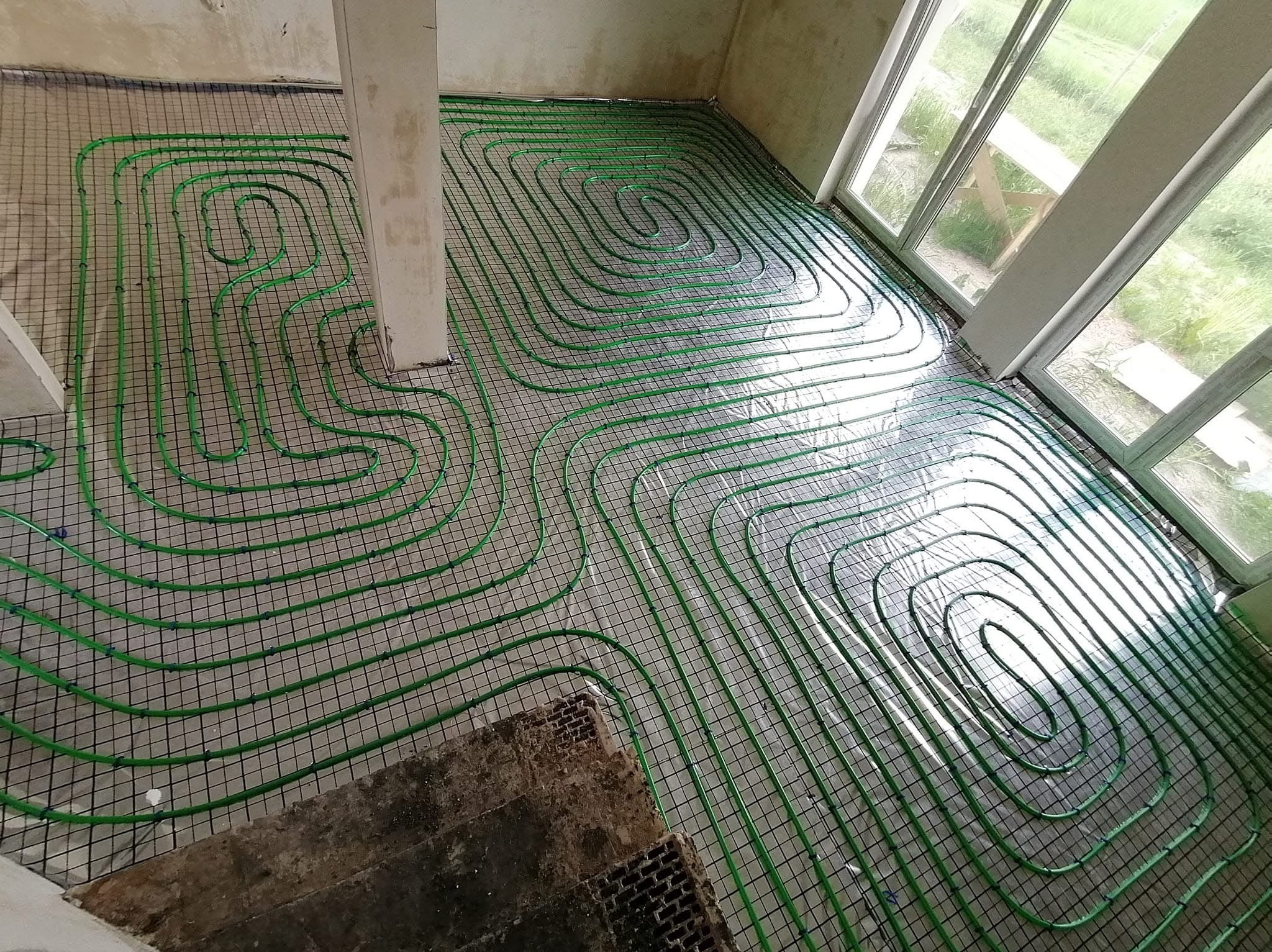 Zielone rurki ogrzewania podłogowego ułożone spiralnie na siatce montażowej w surowym wnętrzu, widoczne okna i fragment starej posadzki.