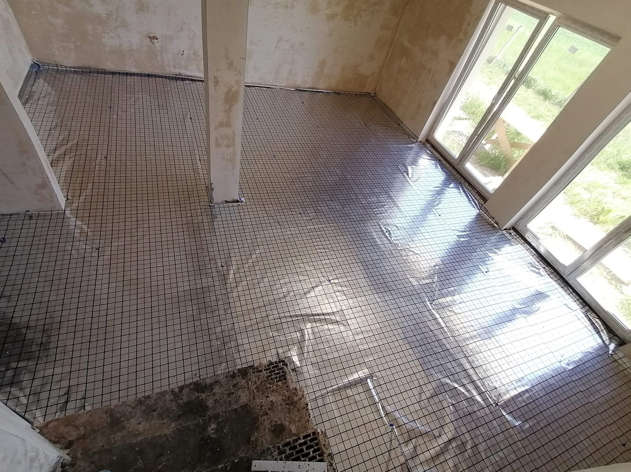 Siatka grzewcza rozłożona na folii w surowym pomieszczeniu z widocznymi fragmentami starej posadzki i kolumną nośną, przygotowana do zalania wylewką.