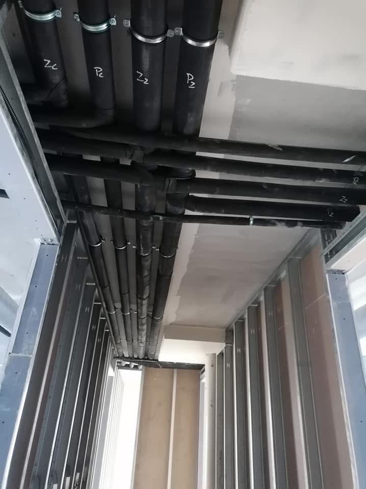 Montaż czarnych rur instalacyjnych z oznaczeniami na suficie w budynku komercyjnym, widoczne metalowe profile ścian działowych.