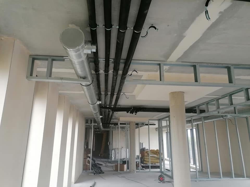 Wnętrze budynku w trakcie montażu instalacji wentylacyjnej z widocznymi czarnymi rurami pod sufitem, srebrnymi kanałami wentylacyjnymi i metalowymi konstrukcjami ścian działowych.