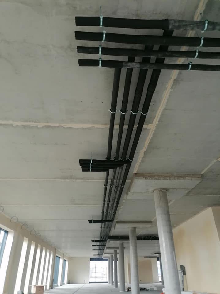 Czarne, zaizolowane rury wentylacyjne przymocowane do sufitu w surowym stanie, widoczne mocowania i fragment wnętrza budynku komercyjnego z kolumnami i oknami.