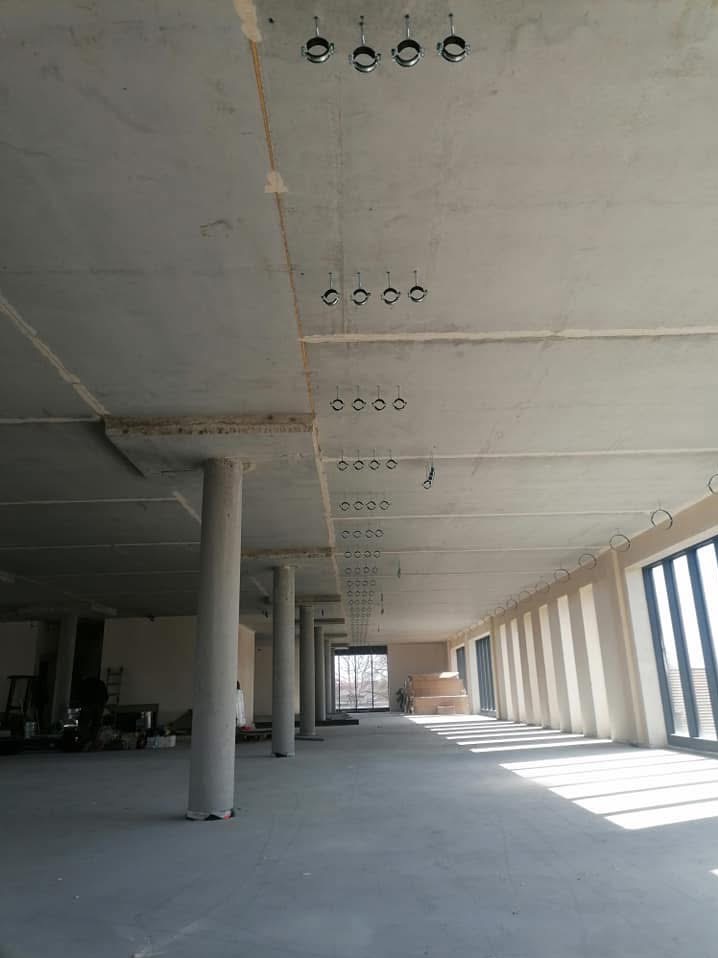 Surowy stan lokalu komercyjnego z widocznymi rzędami uchwytów do instalacji pod sufitem, wspartym na betonowych kolumnach, z promieniami światła wpadającymi przez okna.