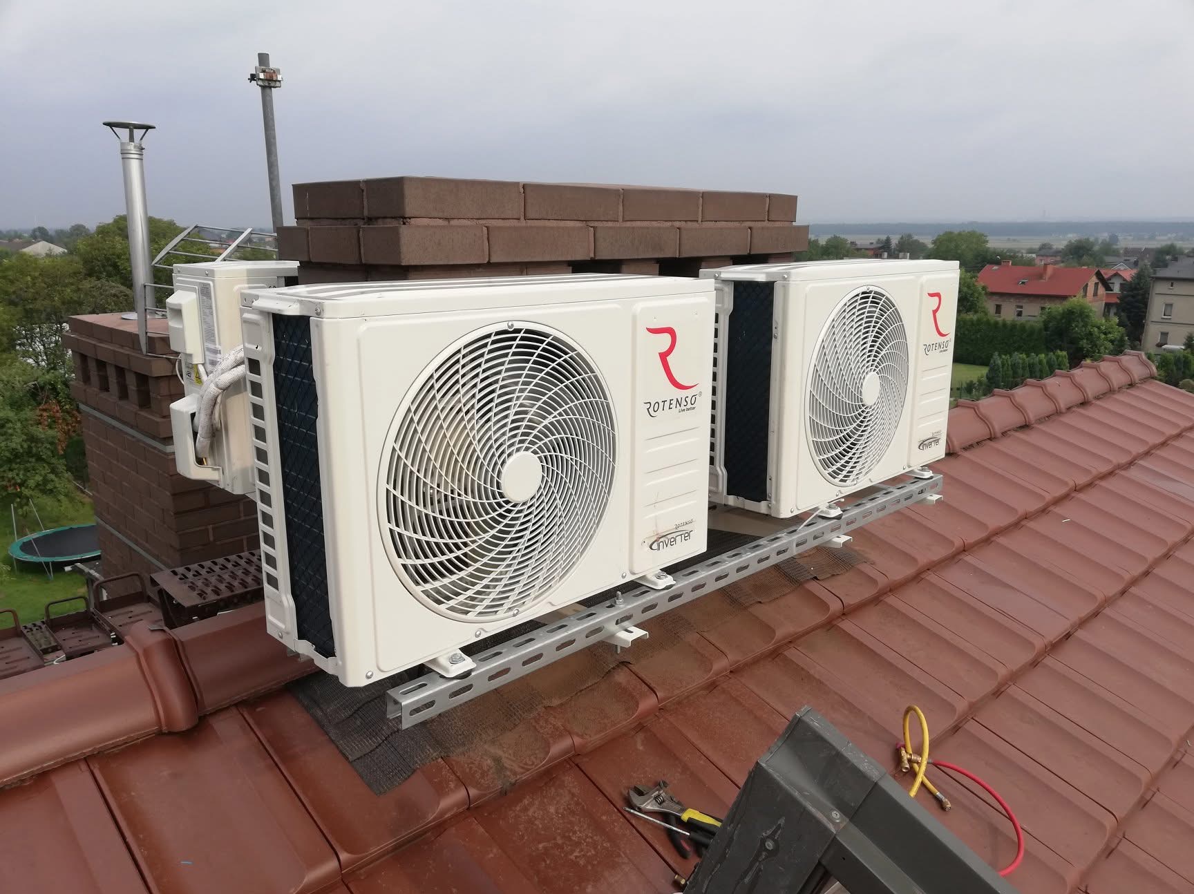 Dwa białe klimatyzatory Rotenso zamontowane na dachu z brązowej dachówki, widok z bliska na instalację, w tle komin i zabudowania.