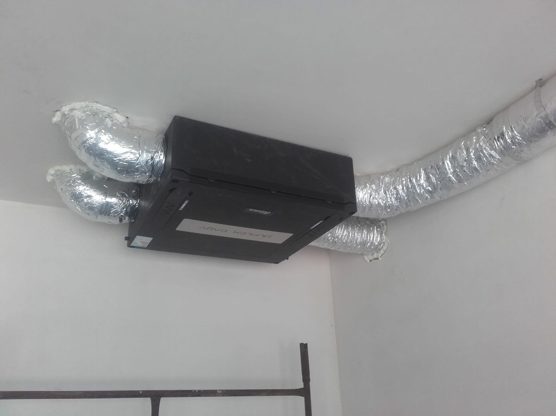 Czarna skrzynka rekuperatora Duplex Easy zamontowana pod sufitem z podłączonymi, izolowanymi przewodami wentylacyjnymi aluminiowymi, widoczny fragment drabiny.