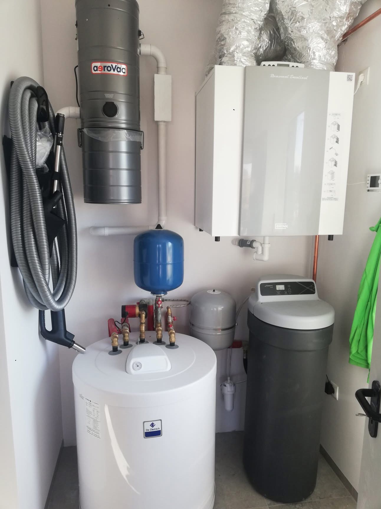Zestaw urządzeń w kotłowni: centralny odkurzacz Aerovac, biały zasobnik ciepłej wody De Dietrich z niebieskim naczyniem przeponowym, szary zmiękczacz wody Trinity oraz centrala wentylacyjna Brink...