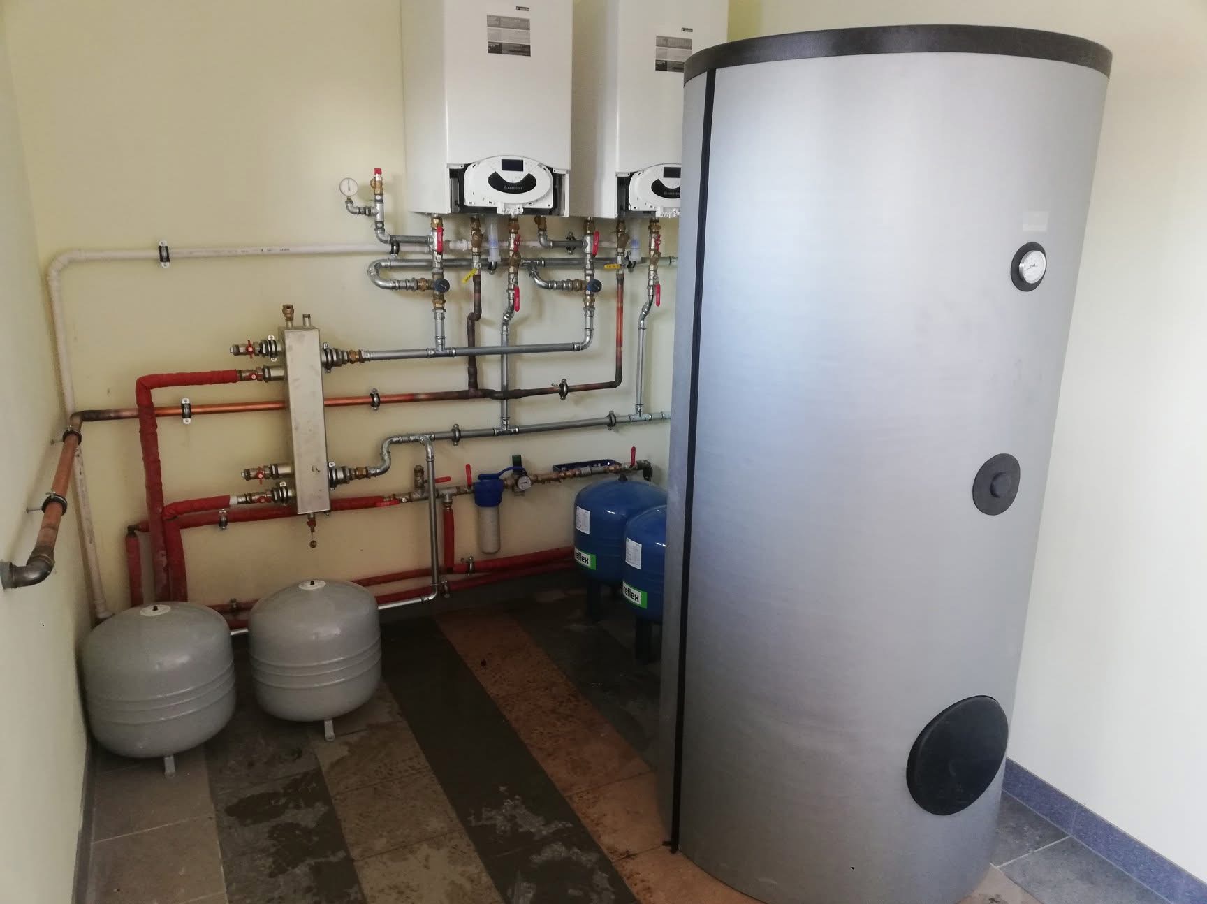 Dwa białe piece grzewcze wiszące na ścianie, połączone rurami miedzianymi i stalowymi, obok duży, szary zbiornik akumulacyjny i dwa niebieskie naczynia przeponowe.