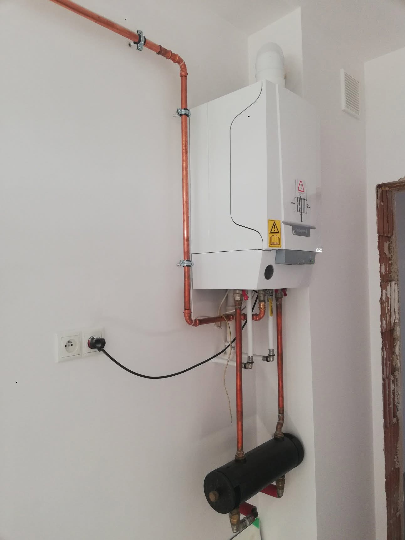 Nowoczesny piec gazowy kondensacyjny, zamontowany na białej ścianie, z widocznymi miedzianymi rurami instalacji grzewczej i czarnym naczyniem przeponowym, podłączony do gniazdka elektrycznego.